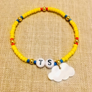 May include: A beaded bracelet featuring a circular strand of small yellow beads interspersed with occasional blue, red, and metallic gold beads. Two white spherical beads with the black letters T and S are centered, accompanied by a white cloud charm.
