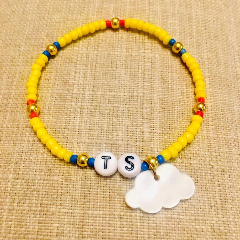 May include: A beaded bracelet featuring a circular strand of small yellow beads interspersed with occasional blue, red, and metallic gold beads. Two white spherical beads with the black letters T and S are centered, accompanied by a white cloud charm.