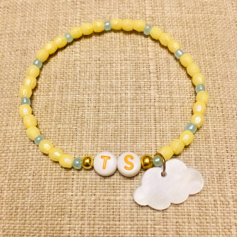 May include: A beaded bracelet featuring yellow faceted beads, small light blue spacer beads, and two white round beads with the orange letters T and S. The bracelet includes two gold-toned metallic beads and a white cloud-shaped charm pendant.