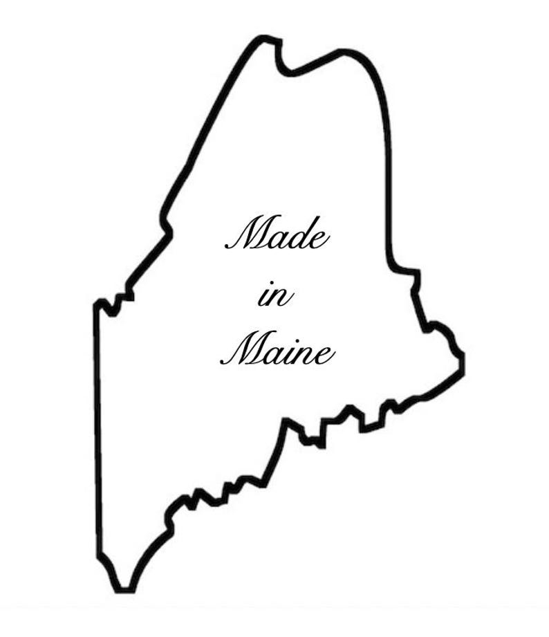 May include: A black outline of the state of Maine on a white background. The words "Made in Maine" are written in a cursive font inside the state outline.