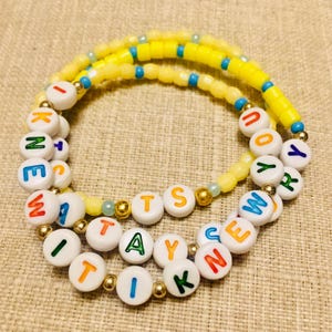May include: A beaded bracelet featuring white circular beads with multicolored letters spelling out I KNEW IT WAS YOU, interspersed with small gold-toned metallic beads and a strand of yellow and blue cylindrical beads arranged in a circular pattern.