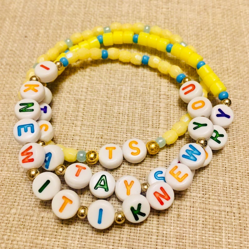 May include: A beaded bracelet featuring white circular beads with multicolored letters spelling out I KNEW IT WAS YOU, interspersed with small gold-toned metallic beads and a strand of yellow and blue cylindrical beads arranged in a circular pattern.