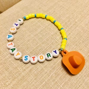 May include: A beaded bracelet featuring yellow cylindrical beads, small blue spacer beads, and white round beads with colorful letters spelling out ITS A TOY STORY. The bracelet includes a single orange cowboy hat charm attached to the side.
