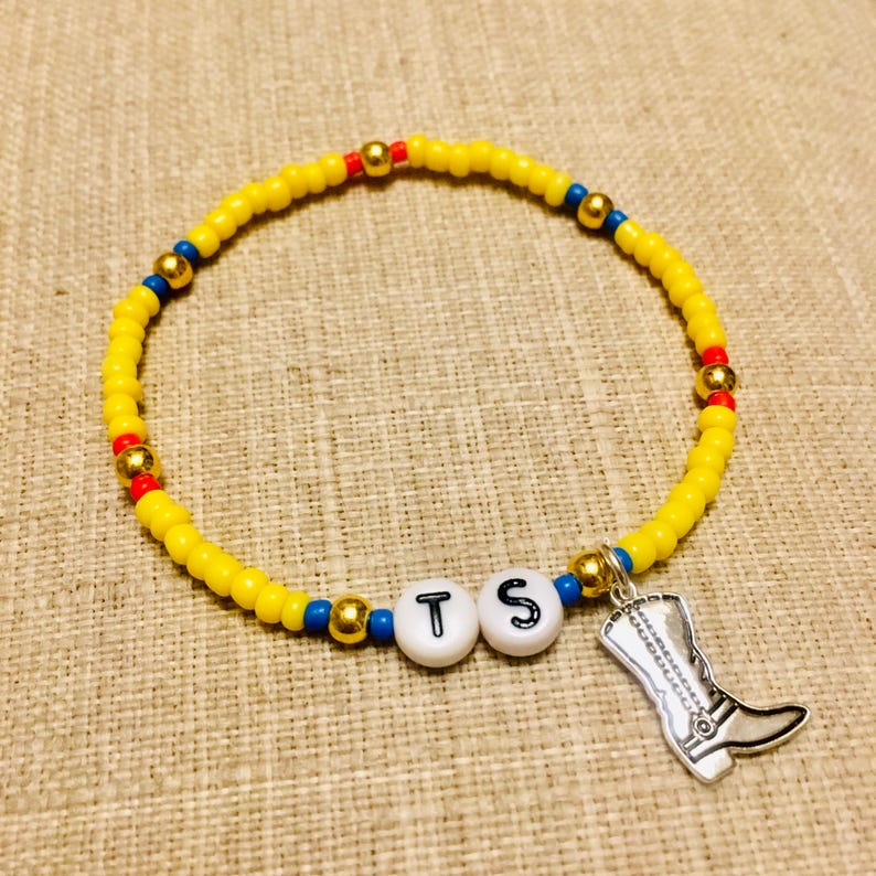 May include: A beaded bracelet featuring small yellow, blue, and red beads interspersed with metallic gold spheres. Two white round beads with the black letters T and S are centered, accompanied by a silver-toned cowboy boot charm pendant.