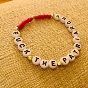 Puede incluir: Una pulsera de cuentas con cuentas redondas blancas con letras negras que forman la frase FUCK THE PATRIARCHY, intercaladas con peque&ntilde;as cuentas met&aacute;licas doradas y un segmento de cuentas rojas cil&iacute;ndricas en dise&ntilde;o circular.