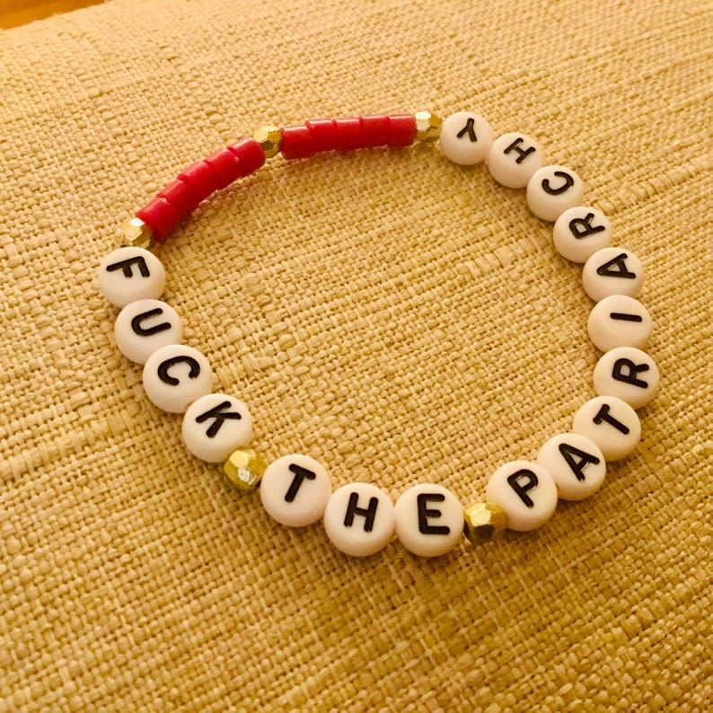 Puede incluir: Una pulsera de cuentas con cuentas redondas blancas con letras negras que forman la frase FUCK THE PATRIARCHY, intercaladas con peque&ntilde;as cuentas met&aacute;licas doradas y un segmento de cuentas rojas cil&iacute;ndricas en dise&ntilde;o circular.