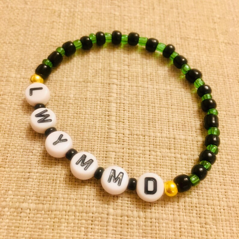 May include: A beaded bracelet with white letter beads spelling out "LWMYMMD". The bracelet features black and green beads, with gold accents. The bracelet is on a neutral-colored surface.