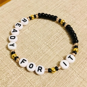 May include: A beaded bracelet with the words "READY FOR IT" spelled out in white letter beads. The bracelet features black, gold, and clear beads, creating a stylish accessory.