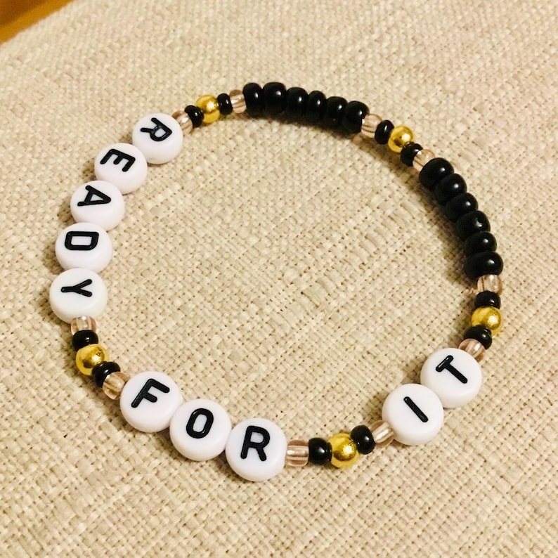 May include: A beaded bracelet with the words "READY FOR IT" spelled out in white letter beads. The bracelet features black, gold, and clear beads, creating a stylish accessory.