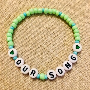 May include: A beaded bracelet with the words "OUR SONG" spelled out in white letter beads. The bracelet features light green and turquoise beads, with heart-shaped accents. The bracelet is on a tan surface.