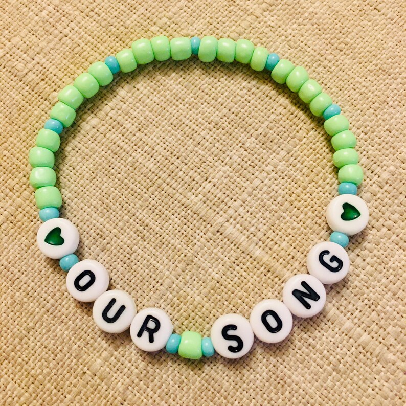 May include: A beaded bracelet with the words "OUR SONG" spelled out in white letter beads. The bracelet features light green and turquoise beads, with heart-shaped accents. The bracelet is on a tan surface.