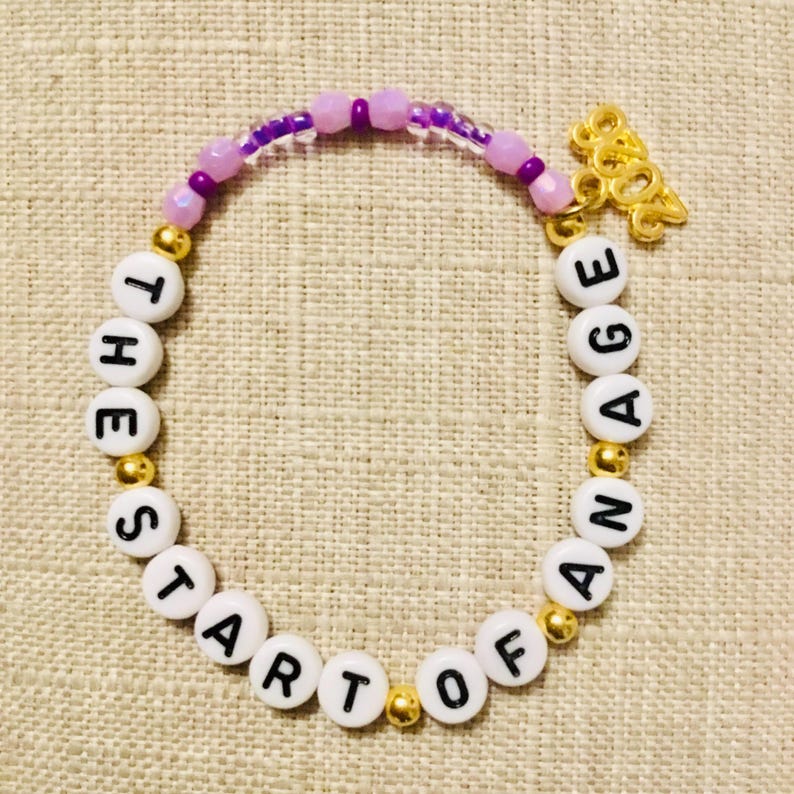 May include: A bracelet with white letter beads spelling "THE START OF AN AGE". The bracelet has gold and purple beads, and a gold charm. The measurements are in centimetres.