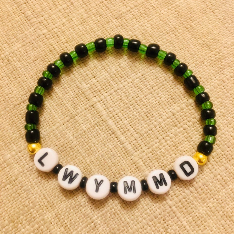 May include: A beaded bracelet with the letters "LWYMMD" spelled out in white letter beads. The bracelet features black and green beads, with gold-colored beads on each end.