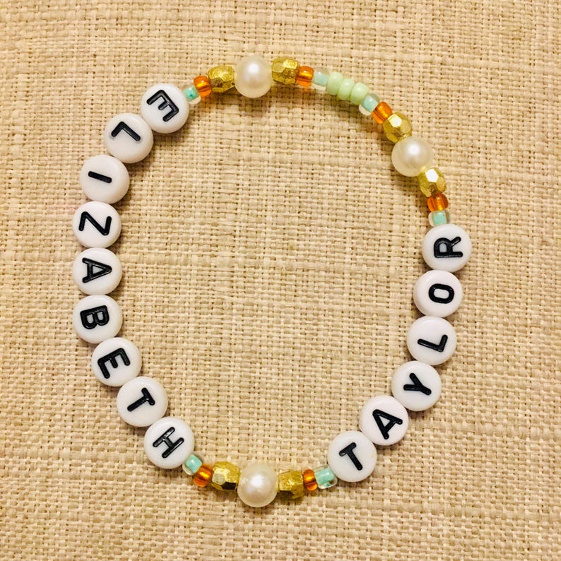 May include: A circular beaded bracelet featuring white round beads with black letters spelling out ELIZABETH TAYLOR. The design includes interspersed gold-toned metallic beads, white faux pearls, and small translucent light green and orange seed beads.