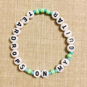 May include: A beaded bracelet with white letter beads spelling "TEAR DROPS ON MY GUITAR". The bracelet features light blue and green accent beads. The bracelet is on a beige surface.