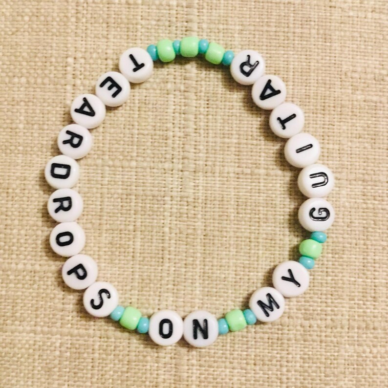 May include: A beaded bracelet with white letter beads spelling "TEAR DROPS ON MY GUITAR". The bracelet features light blue and green accent beads. The bracelet is on a beige surface.