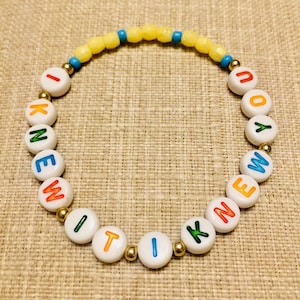 May include: A circular beaded bracelet featuring white round beads with colorful letters spelling I KNEW IT I KNEW YOU, interspersed with small metallic gold beads, and a top section of yellow and blue faceted beads arranged in a repeating pattern.