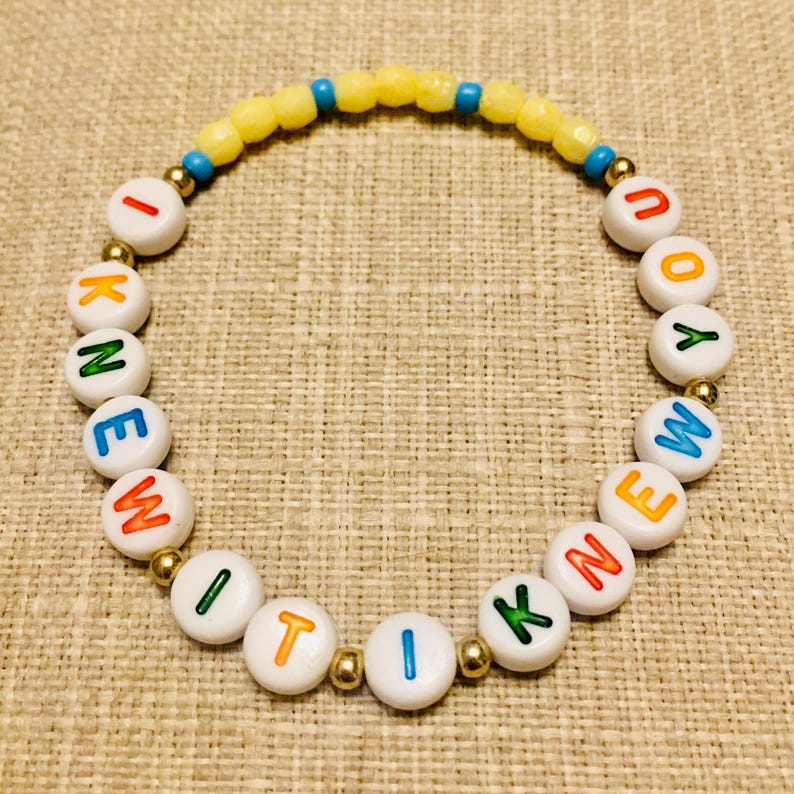 May include: A circular beaded bracelet featuring white round beads with colorful letters spelling I KNEW IT I KNEW YOU, interspersed with small metallic gold beads, and a top section of yellow and blue faceted beads arranged in a repeating pattern.