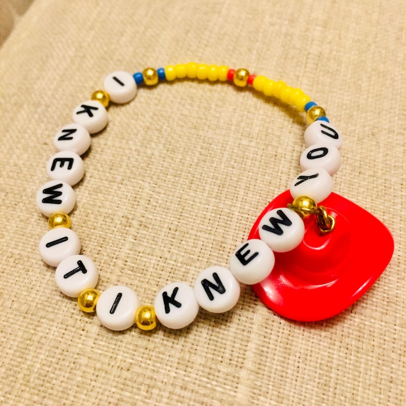 May include: A circular beaded bracelet featuring white round beads with black letters spelling I KNEW IT, interspersed with small metallic gold beads, a section of yellow beads, and a single red cowboy hat charm attached to the bracelet.
