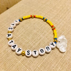 May include: A beaded bracelet featuring white round beads with black letters spelling out ITS A TAY STORY, interspersed with small metallic gold beads, bright yellow beads, and a single white cloud-shaped charm attached to the side of the band.
