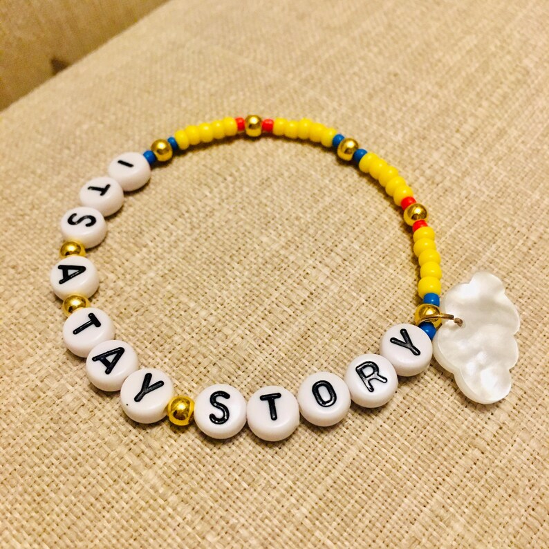 May include: A beaded bracelet featuring white round beads with black letters spelling out ITS A TAY STORY, interspersed with small metallic gold beads, bright yellow beads, and a single white cloud-shaped charm attached to the side of the band.