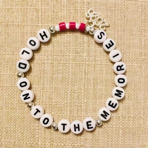 May include: A beaded bracelet with white letter beads spelling "LONDON TO THE MEMORIES". The bracelet has silver accents and a charm that says "2023". The bracelet also has pink and white beads.