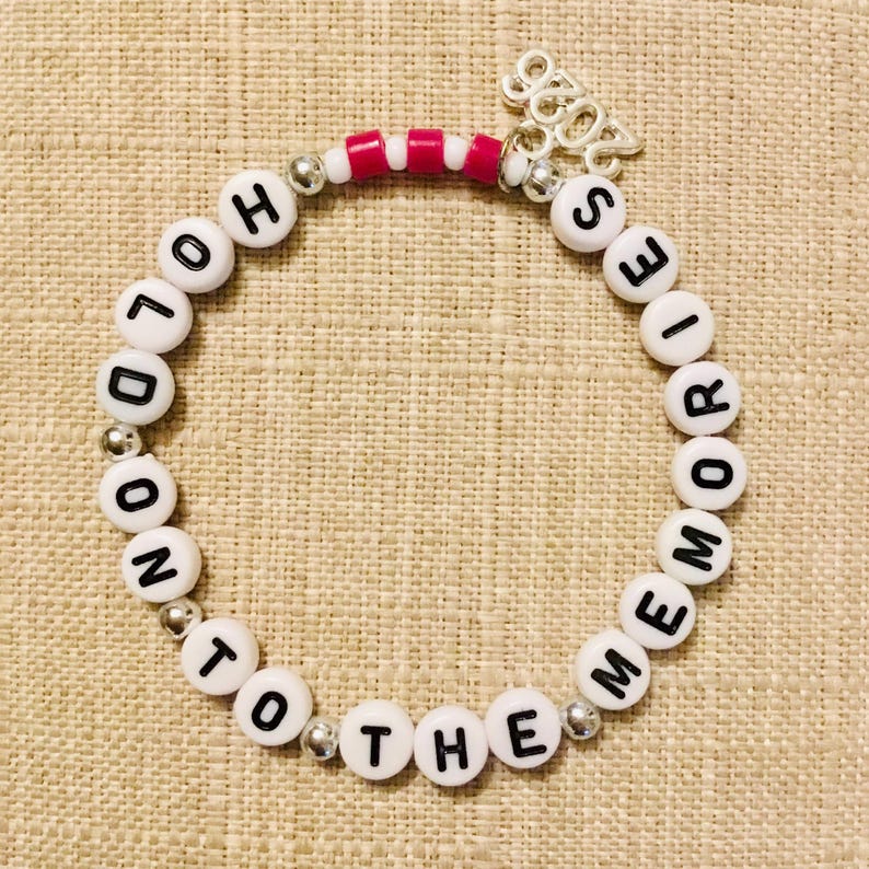 May include: A beaded bracelet with white letter beads spelling "LONDON TO THE MEMORIES". The bracelet has silver accents and a charm that says "2023". The bracelet also has pink and white beads.