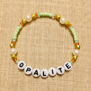 May include: A beaded bracelet with the word "OPALITE" spelled out in white letter beads. The bracelet features a mix of gold, pearl, and green beads. The beads are strung together to form a circular shape.