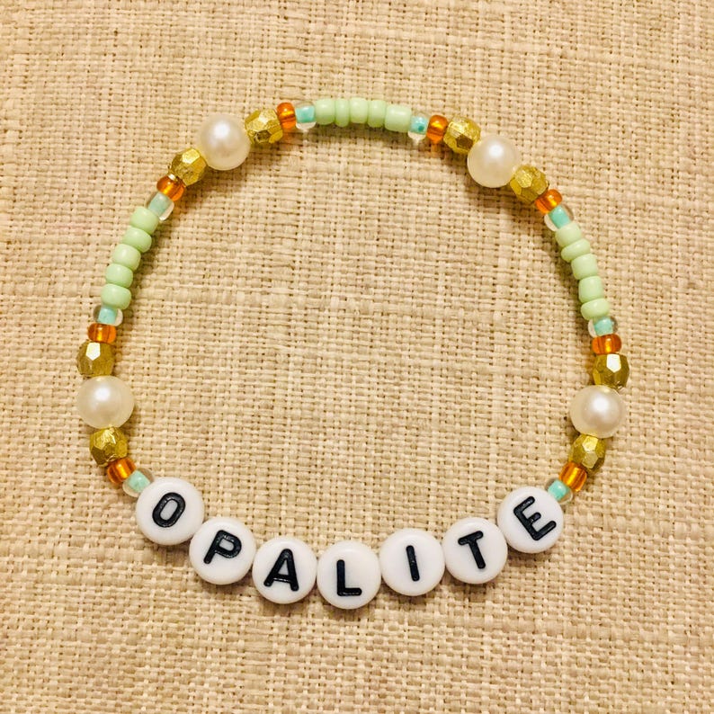 May include: A beaded bracelet with the word "OPALITE" spelled out in white letter beads. The bracelet features a mix of gold, pearl, and green beads. The beads are strung together to form a circular shape.