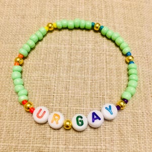 Op de afbeelding: Een cirkelvormige kralenarmband met lichtgroene plastic kralen, afgewisseld met metallic gouden bolvormige kralen en kleine kleurrijke accentkralen. De armband bevat witte letterkralen die de tekst UR GAY vormen.