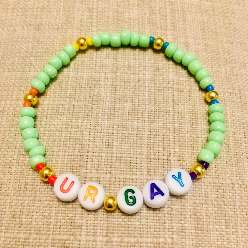 Op de afbeelding: Een cirkelvormige kralenarmband met lichtgroene plastic kralen, afgewisseld met metallic gouden bolvormige kralen en kleine kleurrijke accentkralen. De armband bevat witte letterkralen die de tekst UR GAY vormen.