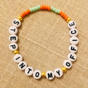 May include: A beaded bracelet with white letter beads spelling "STEP INTO MY OFFICE". The bracelet has orange, green, and gold beads. The bracelet is on a tan surface.