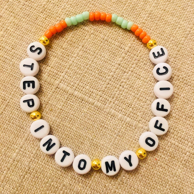 May include: A beaded bracelet with white letter beads spelling "STEP INTO MY OFFICE". The bracelet has orange, green, and gold beads. The bracelet is on a tan surface.
