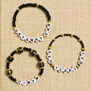 May include: Three beaded bracelets. Two bracelets have black beads with gold accents and white letter beads spelling "GETAWAY CAR" and "NEW YEARS DAY". The third bracelet has black beads, gold accents, and iridescent beads.