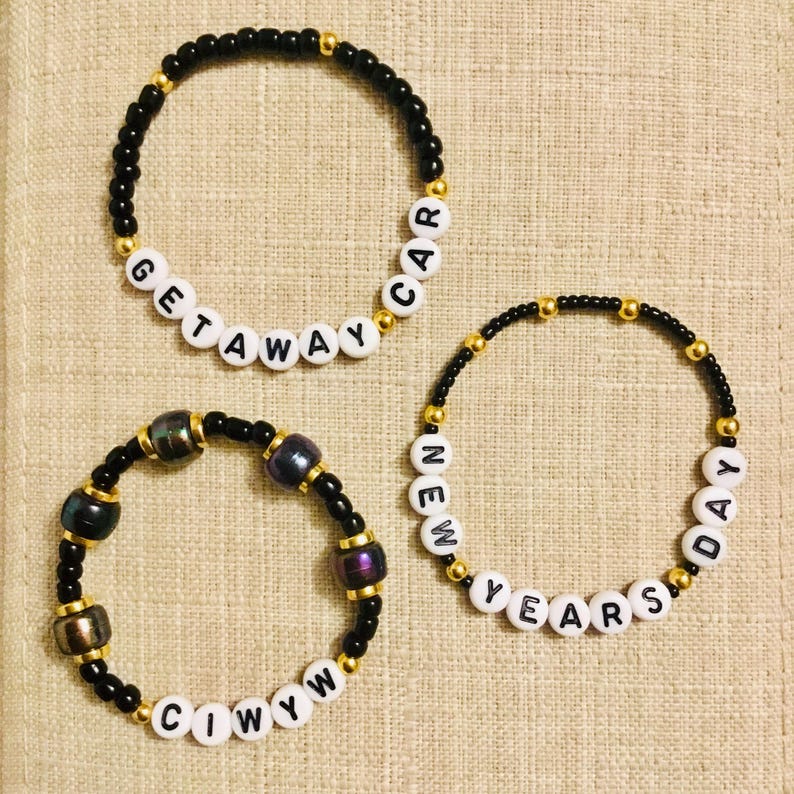 May include: Three beaded bracelets. Two bracelets have black beads with gold accents and white letter beads spelling "GETAWAY CAR" and "NEW YEARS DAY". The third bracelet has black beads, gold accents, and iridescent beads.