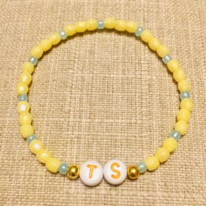 May include: A circular beaded bracelet featuring alternating yellow and light blue beads. Two white spherical beads with the orange letters T and S are positioned at the bottom, flanked by two small metallic gold beads on either side of the letters.