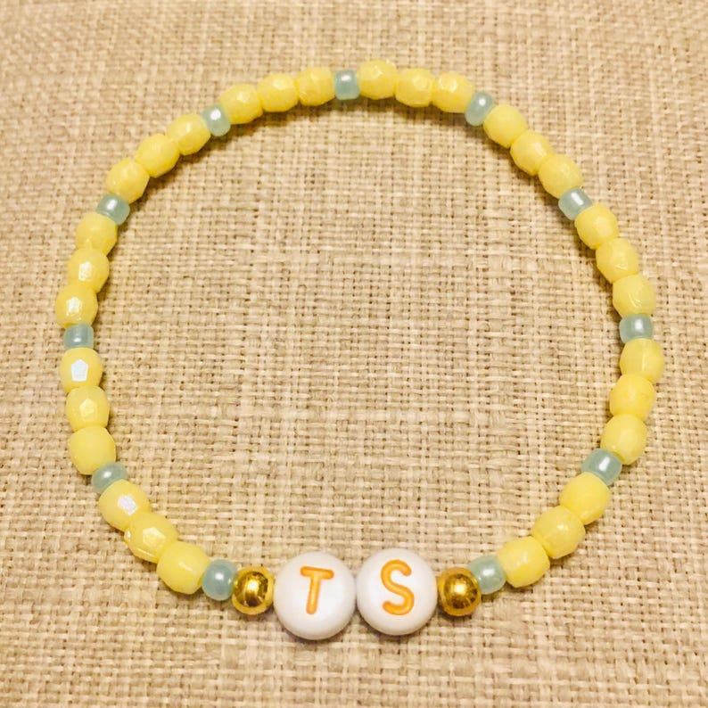 May include: A circular beaded bracelet featuring alternating yellow and light blue beads. Two white spherical beads with the orange letters T and S are positioned at the bottom, flanked by two small metallic gold beads on either side of the letters.