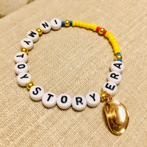 May include: A beaded bracelet featuring white round beads with black letters spelling out IN MY TOY STORY ERA, interspersed with metallic gold beads, a section of yellow seed beads, and a dangling metallic gold cowboy hat charm.