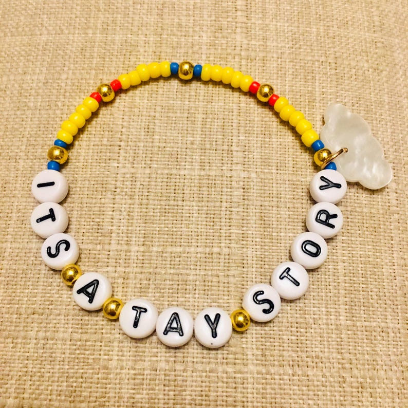 May include: A beaded bracelet featuring yellow, red, and blue seed beads interspersed with gold-tone metallic spheres. White circular beads with black lettering spell out ITS A TAY STORY, accompanied by a translucent white cloud-shaped charm.