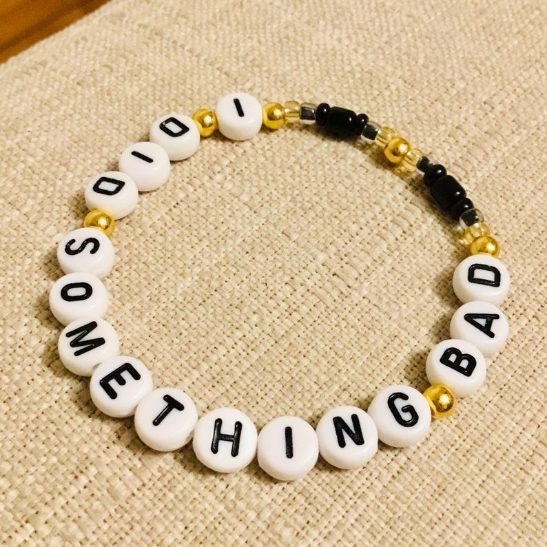 May include: A beaded bracelet with white letter beads spelling "SOMETHING BAD". The bracelet features gold and black accent beads. The bracelet is on a beige surface.