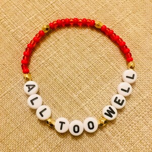 Puede incluir: Una pulsera de cuentas circular que presenta peque&ntilde;as cuentas rojas transl&uacute;cidas, cuentas espaciadoras doradas facetadas y cuentas alfab&eacute;ticas blancas redondas con letras negras que deletrean la frase ALL TOO WELL en una secuencia continua.