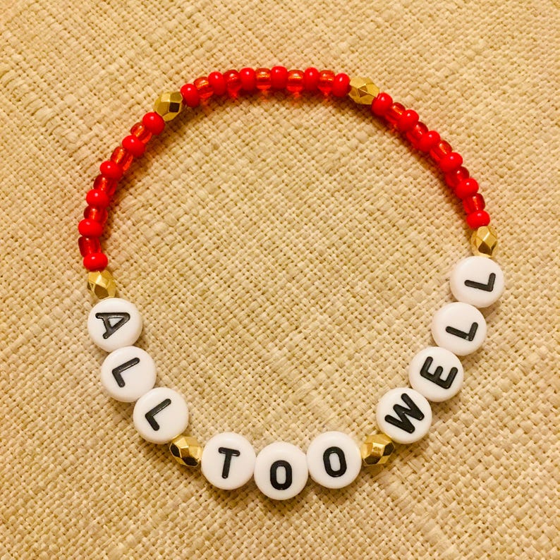 Puede incluir: Una pulsera de cuentas circular que presenta peque&ntilde;as cuentas rojas transl&uacute;cidas, cuentas espaciadoras doradas facetadas y cuentas alfab&eacute;ticas blancas redondas con letras negras que deletrean la frase ALL TOO WELL en una secuencia continua.