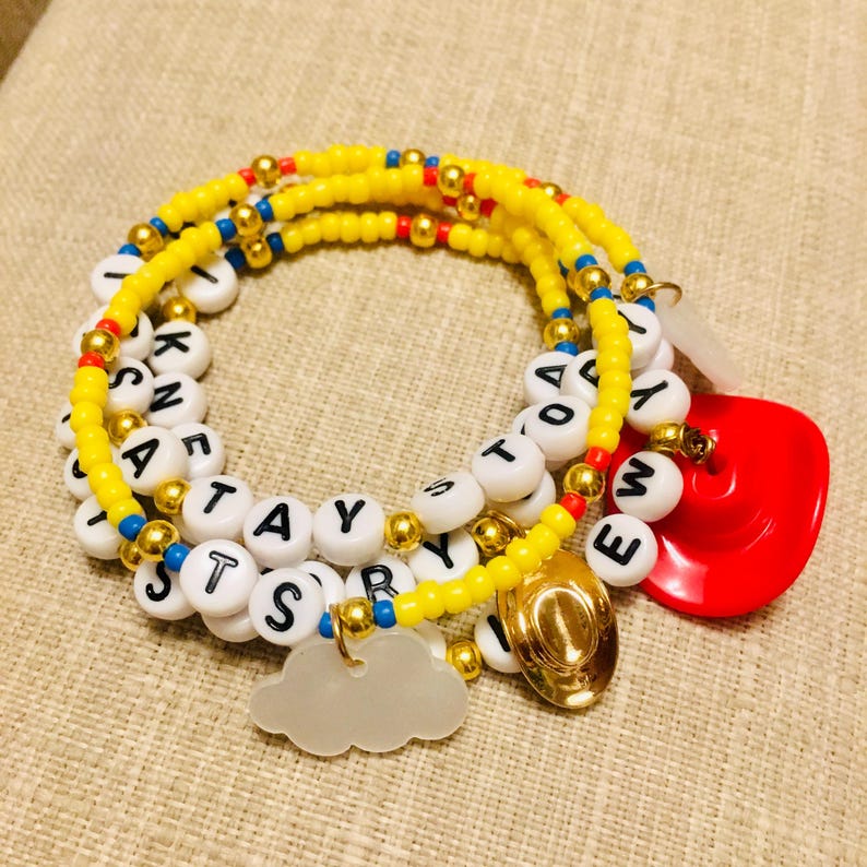 May include: A multi-strand beaded bracelet featuring yellow, red, and blue seed beads, gold-tone spacer beads, and white letter beads spelling TOY STORY. The bracelet includes a red cowboy hat charm, a gold cowboy hat charm, and a white cloud charm.