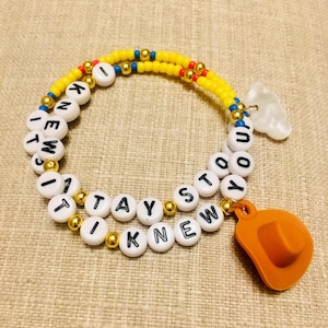 May include: A beaded bracelet featuring yellow, blue, and metallic gold beads interspersed with white letter beads spelling out I KNEW IT STAYED I KNEW IT. The bracelet includes a white cloud charm and an orange cowboy hat charm attached.