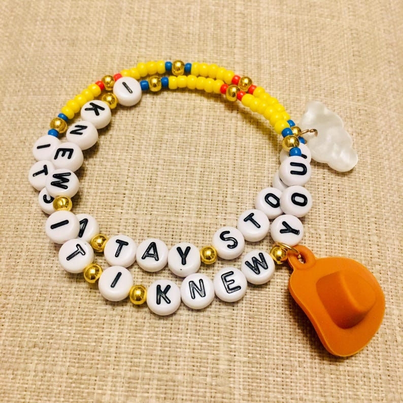 May include: A beaded bracelet featuring yellow, blue, and metallic gold beads interspersed with white letter beads spelling out I KNEW IT STAYED I KNEW IT. The bracelet includes a white cloud charm and an orange cowboy hat charm attached.