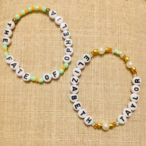 May include: Two beaded bracelets. One bracelet spells "THE FATE OF POPELIA" in white letter beads, accented with green and orange beads. The other spells "ELIZABETH TAYLOR" with white letter beads, gold and pearl accents.