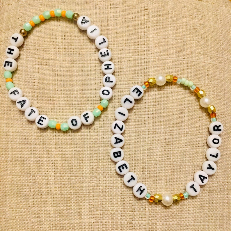 May include: Two beaded bracelets. One bracelet spells "THE FATE OF POPELIA" in white letter beads, accented with green and orange beads. The other spells "ELIZABETH TAYLOR" with white letter beads, gold and pearl accents.