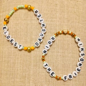 May include: Two beaded bracelets. One bracelet spells "ELDEST DAUGHTER" in white letter beads, accented with gold and orange beads. The other bracelet spells "THE FRIENDSHIP RUINING" with similar beads and a gold heart charm.