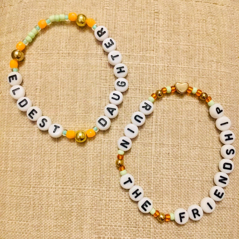 May include: Two beaded bracelets. One bracelet spells "ELDEST DAUGHTER" in white letter beads, accented with gold and orange beads. The other bracelet spells "THE FRIENDSHIP RUINING" with similar beads and a gold heart charm.