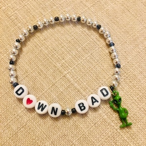 May include: A beaded bracelet with silver and black beads. The bracelet spells out "DOWN BAD" with white letter beads and a red heart. A green alien charm hangs from the bracelet.